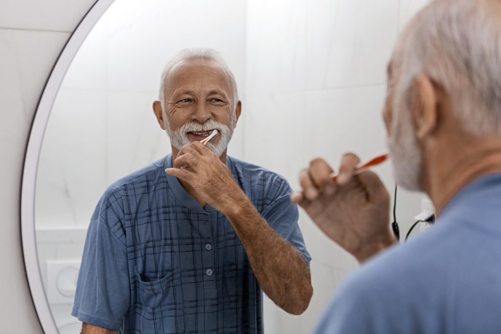 Senior man brushing his teeth looking in bathroom mirror